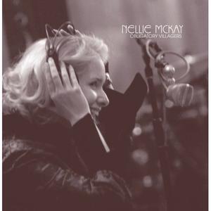 Couverture de l’album “Obligatory Villagers” de Nellie McKay. C’est une photo en noir et blanc de la chanteuse face à un micro. Elle à les mains sur le casque audio posé sur sa tête et semble rire.