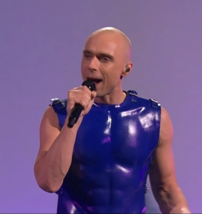 Capture d'écran du candidat Letton à l'Eurovision, dans un tanktop de cuir bleu qui souligne (ou ajoute?) des pecs et des abdos de ouf au gugusse